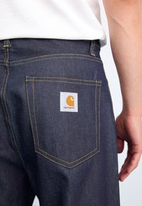 Dark denim jeans featuring yellow stitching, a back pocket label with the Carhartt logo, and a standard fit outline.
