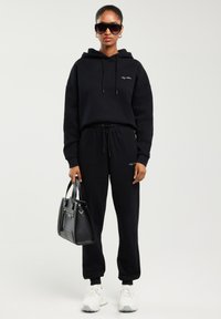 Žena v černé mikině s kapucí a teplácích, bílých teniskách, velkých černých slunečních brýlích, držící černou kabelku na jednobarevném pozadí.