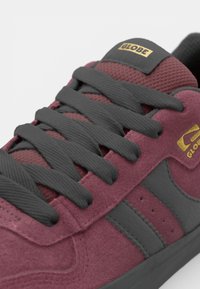 Burgundy suede sneaker with grey leather accents, black laces, and a golden logo patch on the tongue. Textured upper with stitching details.