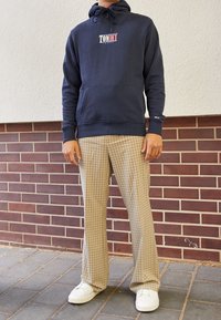 Navy blue hoodie with "TOMMY" logo, paired with beige plaid pants and white sneakers. Standard fit and soft fabric.