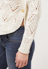 Stickad kofta i krämfärg med ett öppet mönster, med ribbad nederkant och ärmslut. En enda träknapp pryder framsidan.