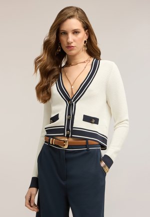 Mujer con cabello largo y ondulado que lleva un cárdigan corto blanco con ribete negro, pantalones azul marino, cinturón marrón, pendientes dorados, collares en capas y pulseras.