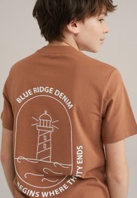 Ruda medvilnės marškinėliai su švyturio grafika balta spalva ir užrašais "Blue Ridge Denim" bei "Pradžia ten, kur baigiasi išpardavimas" aplink jį.