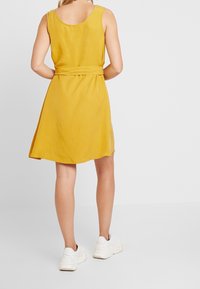 Mosterdgeel mouwloos jurkje met een ceintuur in de taille, vloeiende rok en ronde halslijn, gecombineerd met witte sneakers. Glad textuur.