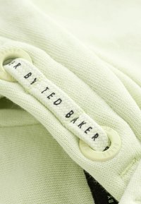 Kratki prikaz kremastog materijala s tkanom vezicom provučenom kroz plastične okvire, s natpisom "TED BAKER" ispisanim crnim slovima.