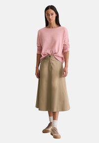 Lichtroze trui met brede halslijn, ingestopt in een knielange tan rokje met een knoopdetail, gecombineerd met beige sneakers en witte sokken.