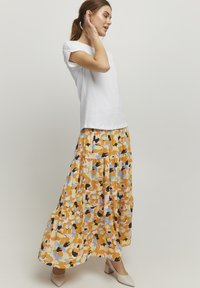 Witte shirt met korte mouwen gecombineerd met een lange, gelaagde rok met een bloemenpatroon in geel, oranje en blauw, gedragen met beige hakken.