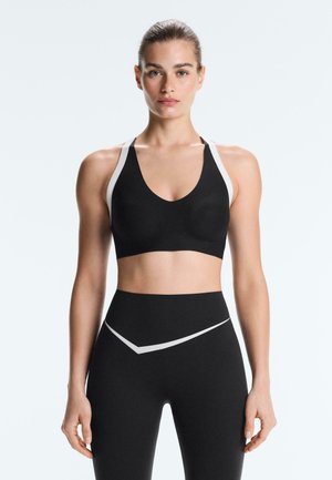 Młoda kobieta stoi, ubrana w czarny sportowy biustonosz i legginsy z wysokim stanem z białymi paskami na ramionach i biodrach.