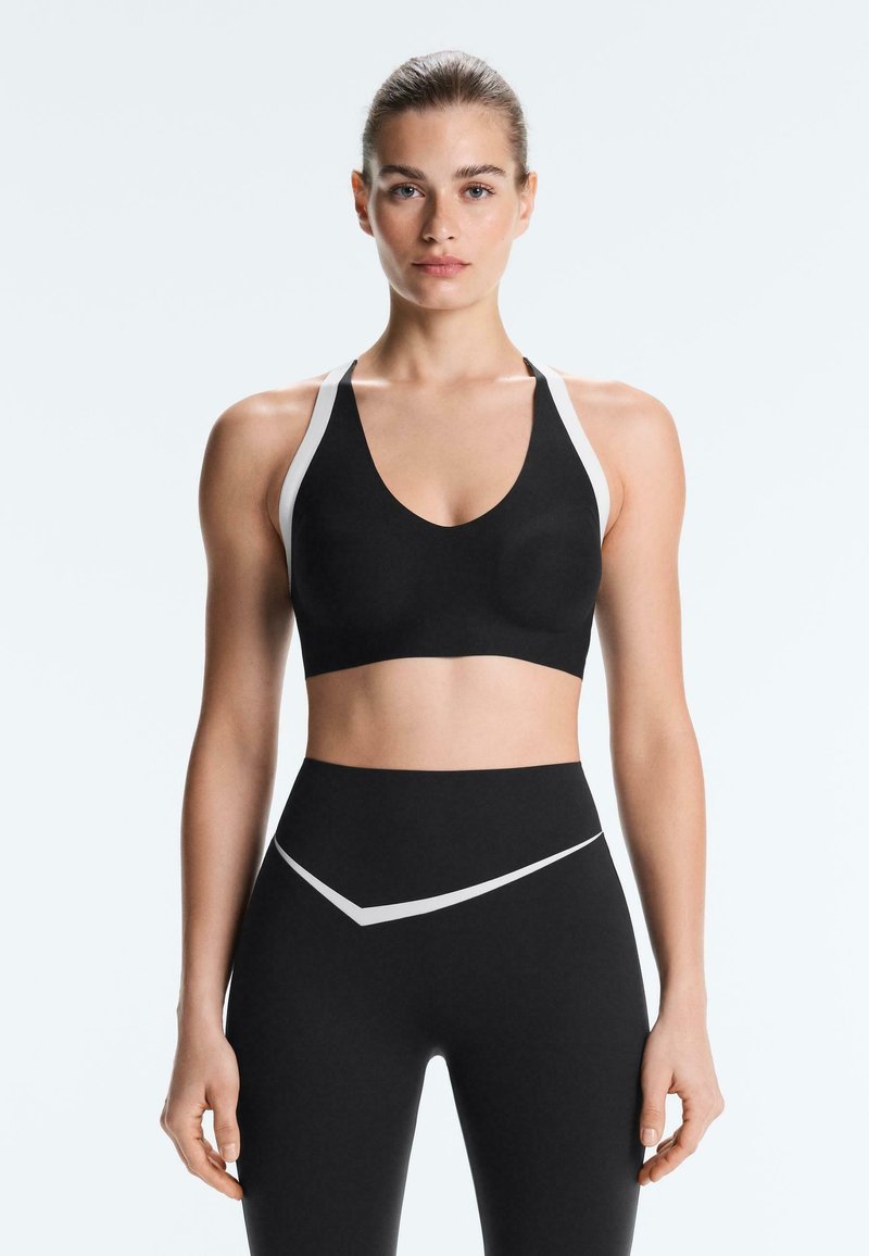 Jeune femme debout, portant un soutien-gorge de sport noir et des leggings taille haute avec des bandes blanches sur les épaules et les hanches.