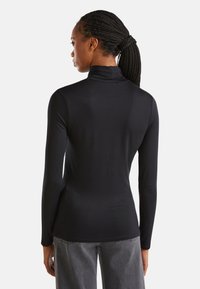 Donna con i capelli intrecciati in un ponytail, che indossa un maglione nero a maniche lunghe e jeans grigi, vista di spalle su uno sfondo bianco.
