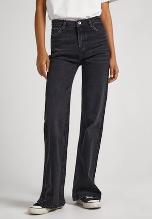 Personne portant un jean noir à jambes larges associé à des baskets noires et blanches, debout devant un fond uni clair.