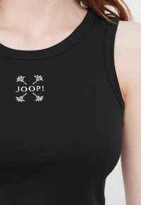 Débardeur noir à côtes avec un motif brodé blanc représentant des fleurs et le texte "JOOP !" au centre. Encolure ronde et sans manches.