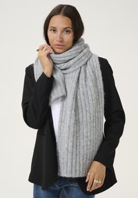 Écharpe grise en laine douce avec des côtes, enroulée autour du cou. Portée par-dessus un blazer noir, mettant en valeur la texture et la chaleur.
