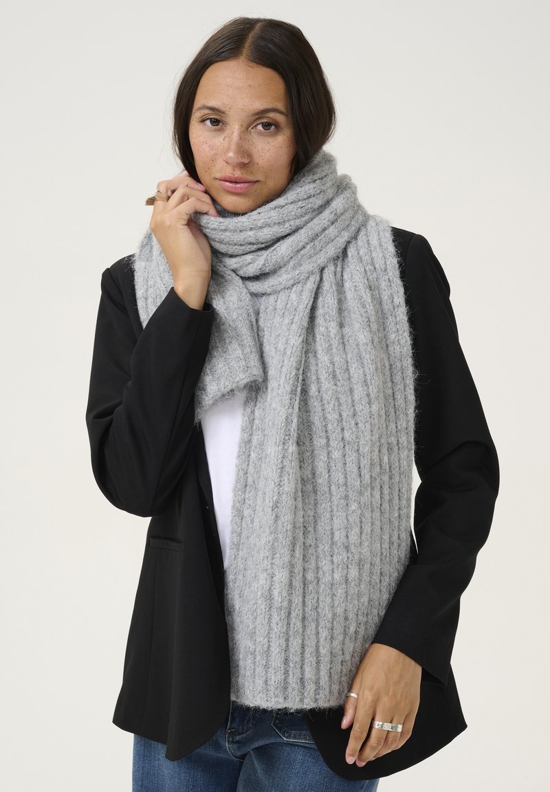 Écharpe grise en laine douce avec des côtes, enroulée autour du cou. Portée par-dessus un blazer noir, mettant en valeur la texture et la chaleur.