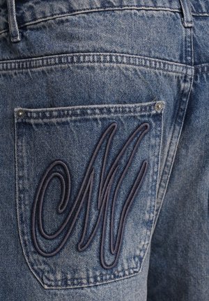 Denim jeans featuring a dark blue embroidered "M" on the back pocket, with a classic five-pocket design and medium blue wash texture.