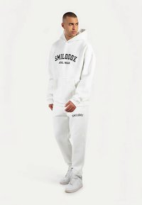 Weißes Kapuzenpullover- und Jogginghosen-Set mit einer Fronttasche, schwarzer Aufschrift "SMILODOX ATHL WEAR" und weißen Turnschuhen. Glatte Textur, lässiges Design.