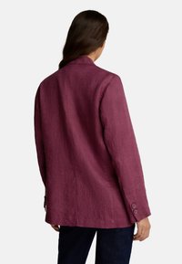 Femme avec de longs cheveux bruns portant un blazer ample prune et un pantalon sombre, vue de dos sur un fond uni.
