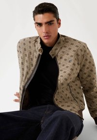 Veste bomber beige à motif avec fermeture éclair, présentant un design géométrique et une doublure foncée. Portée sur une chemise noire, assortie à un jean foncé.