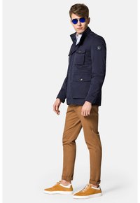 Granatowa wodoodporna kurtka z wieloma kieszeniami, wysokim kołnierzem i zapięciem na guziki, połączona z jasnobrązowymi chinosami i żółtymi sneakersami.