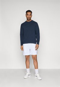 Felpa navy con collo a giro e dettagli testuali, abbinata a pantaloni corti bianchi. Scarpe da ginnastica e calze bianche completano il look sportivo.
