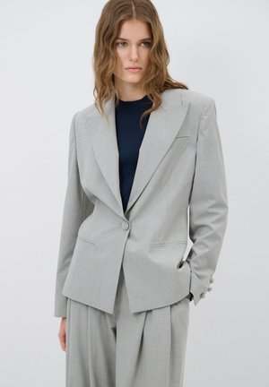 Blazer grigio chiaro con un design strutturato, chiusura a un bottone e due tasche frontali. Abbinato a un top blu scuro e pantaloni larghi.