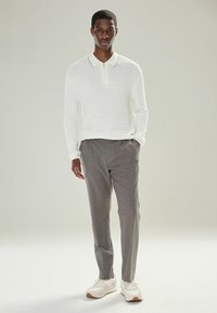 Jeune homme se tenant les mains dans les poches, portant une chemise à manches longues zippée texturée blanche, un pantalon gris et des baskets blanches.