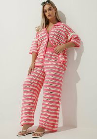 Różowy dzianinowy zestaw wypoczynkowy składający się z kardiganu z krótkim rękawem oraz spodni o szerokich nogawkach z kontrastującymi białymi paskami i haftem.