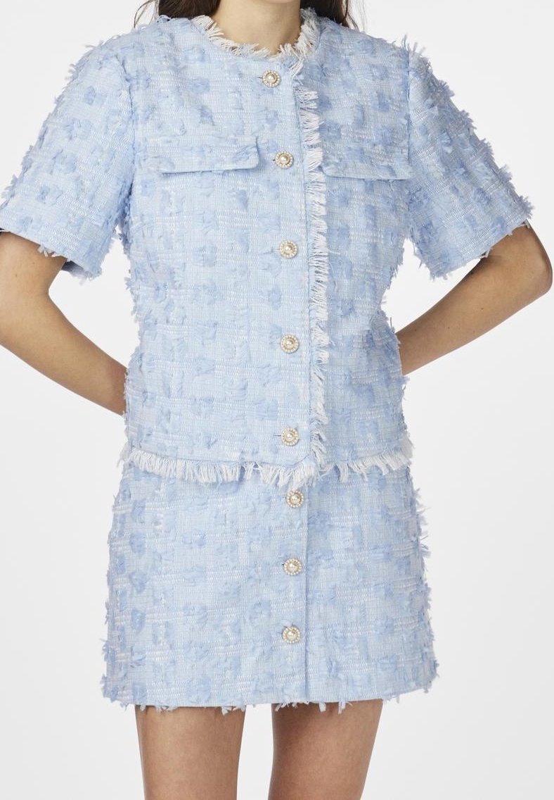 Veste courte à manches texturée bleu clair et jupe assortie avec des boutons en perles et des bords frangés, portée par une personne avec les mains dans le dos.