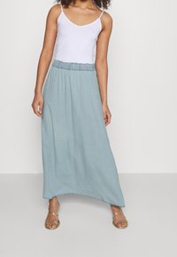 Kvinna som bär en vit ärmlös topp och en ljusblå ankel-lång kjol med resårmidja, stående med guld sandaler.