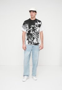 Μπλουζάκι με κοντά μανίκια σε ασπρόμαυρο tie-dye, συνδυασμένο με ανοιχτό μπλε, χαλαρά τζιν και λευκά αθλητικά παπούτσια. Απλό λευκό καπέλο στο κεφάλι.