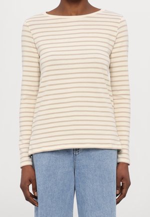 Personne portant un haut à manches longues à rayures horizontales crème et beige, associé à un jean en denim bleu clair, debout devant un fond uni.