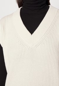 Strick-Weste in Creme mit einem V-Ausschnitt; strukturiertes Rippmuster; kombiniert mit einem schwarzen Rollkragenpullover darunter.