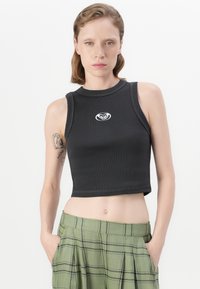 Young woman with short brown hair wears black cropped tank top with logo and green plaid pants, hands in pockets, neutral expression.