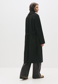 Čierny kabát z vlnenej zmesi so širokým golierom, opaskom v páse a dlhými rukávmi, skombinovaný so širokými tmavými džínsami a hnedými členkovými čižmami.