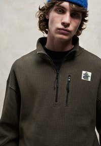 Hnedý flísový pulóver s textúrou a zipsom na krku, ozdobený malou logovou nášivkou a čiernymi detailmi na zipsu. Nosený cez čierne tričko.