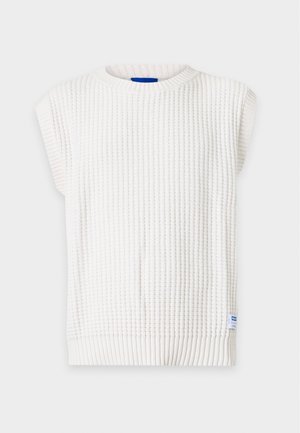 Homme portant une chemise à carreaux beige avec les manches retroussées, un pull sans manches en tricot blanc, un short en jean clair, des chaussettes noires et des chaussures noires.