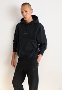 Sort hættebluse med kangaroo-lomme og snørelukning. Sammen med matchende sorte sweatpants med et lille logo-patch.