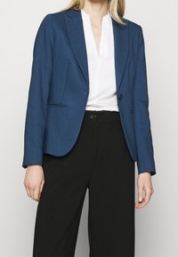 Blazer azul marino con un corte ajustado, cierre de un solo botón, solapas muesca y dos bolsillos frontales. Lleva encima una blusa blanca y pantalones negros.
