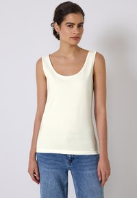 Débardeur crème à encolure arrondie, sans manches, avec une texture lisse et douce. Accompagné de jeans en denim bleu.