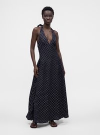 Femme portant une robe longue noire dos nu à pois blancs et des sandales noires, debout devant un fond uni.