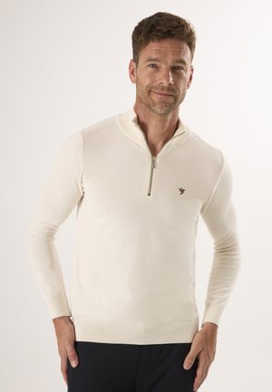 Crème kleurige pullover trui met een halve rits. Heeft ribgebreide manchetten en onderkant, en een klein bruin logo aan de bovenste linkerkant. Glad textuur.