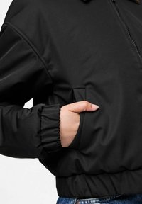 Ręka włożona w boczną kieszeń czarnej kurtki z zamkiem błyskawicznym, z elastycznymi mankietami i pasem, noszonej na niebieskich dżinsach.