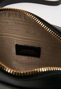 Interior negro con cremallera de un bolso que presenta un parche con el logotipo, cremallera dorada y un forro beige con un patrón que repite "LAUREN RALPH LAUREN".