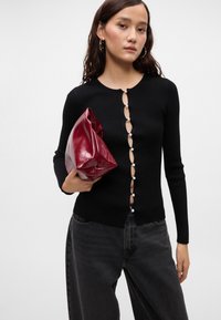 Črna rebrasta jakna z bisernimi gumbi spredaj, v kombinaciji z sijočo burgundsko torbico in temno denim hlačami.