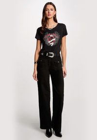 T-shirt grafico nero con un design di rosa e ali con testo "Rock & Roll". Abbinato a pantaloni neri a vita alta e una cintura argentata.