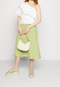 Top bianco a spalla singola con dettagli a balza, gonna midi plissettata verde, borsa di colore chiaro e sandali con piattaforma con accenti floreali.