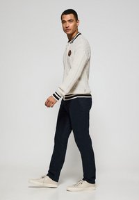 Polo blanco de punto trenzado con cuello a rayas en blanco y negro, pantalones azul marino y zapatillas beige. Presenta un logo en el pecho.