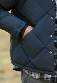 Jachetă din material matlasat de culoare bleumarin, cu închidere prin capse, având un buzunar lateral. Pur și simplu peste o cămașă în carouri, scoțând în evidență o textură netedă și un model în romb.