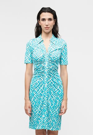 Γυναίκα με σκούρα μαλλιά που φοράει ένα γαλάζιο φόρεμα με ραφές και κουμπιά, με λευκό γεωμετρικό μοτίβο, στέκεται μπροστά σε απλό γκρίζο φόντο.