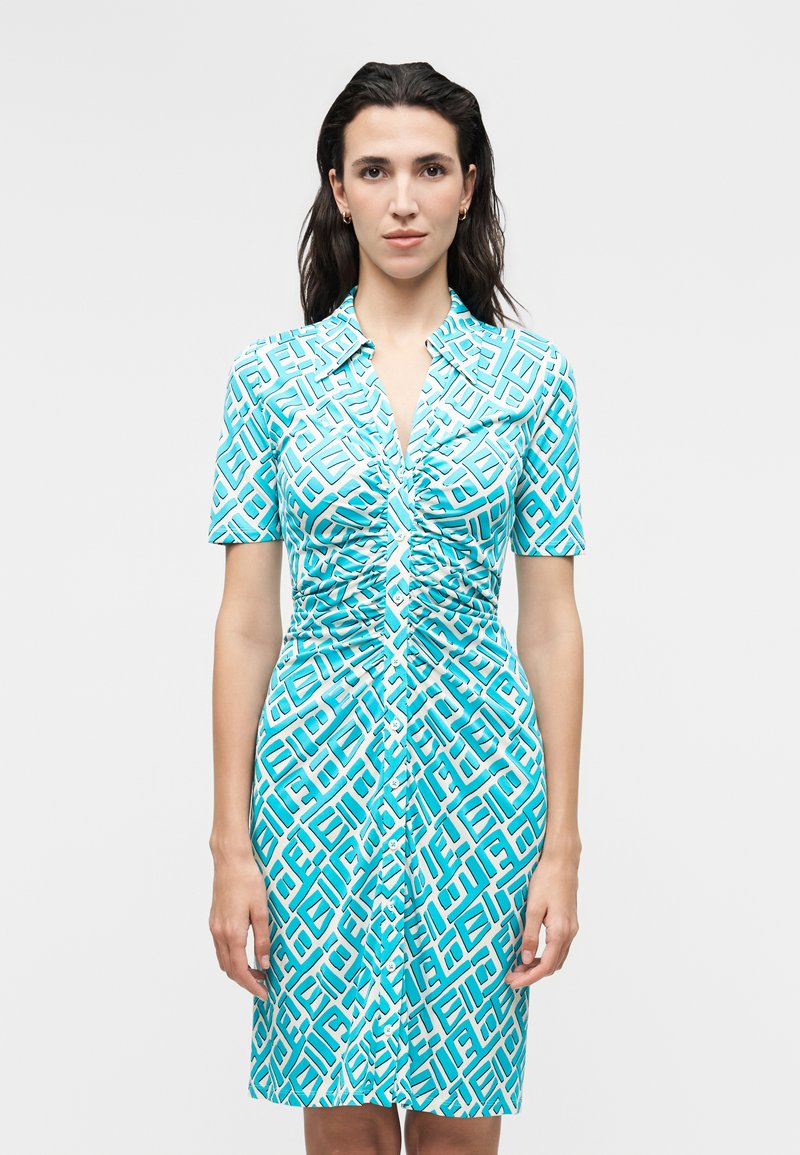 Γυναίκα με σκούρα μαλλιά που φοράει ένα γαλάζιο φόρεμα με ραφές και κουμπιά, με λευκό γεωμετρικό μοτίβο, στέκεται μπροστά σε απλό γκρίζο φόντο.